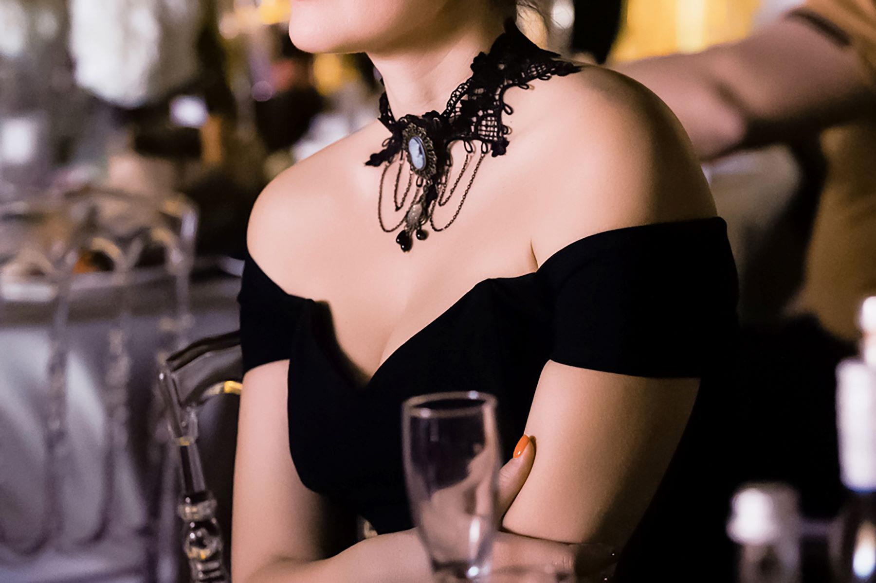 Elegant woman in a black off-shoulder dress with a decorative choker, seated at a luxurious venue with drinks nearby.