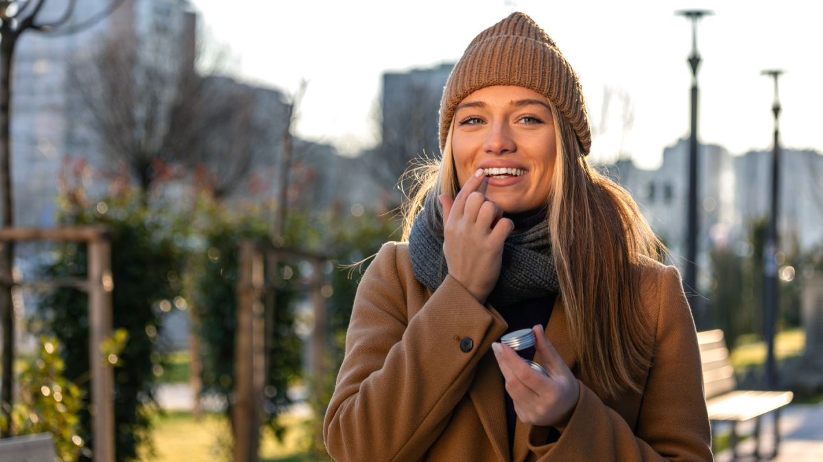 Woman applying lip balm outdoors, smiling, wearing a knit hat and coat on a sunny day.