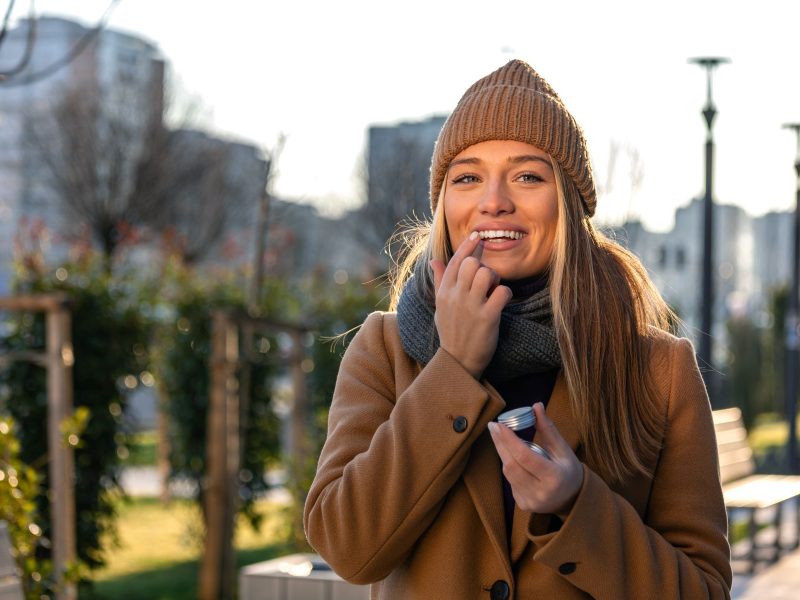 Woman applying lip balm outdoors, smiling, wearing a knit hat and coat on a sunny day.