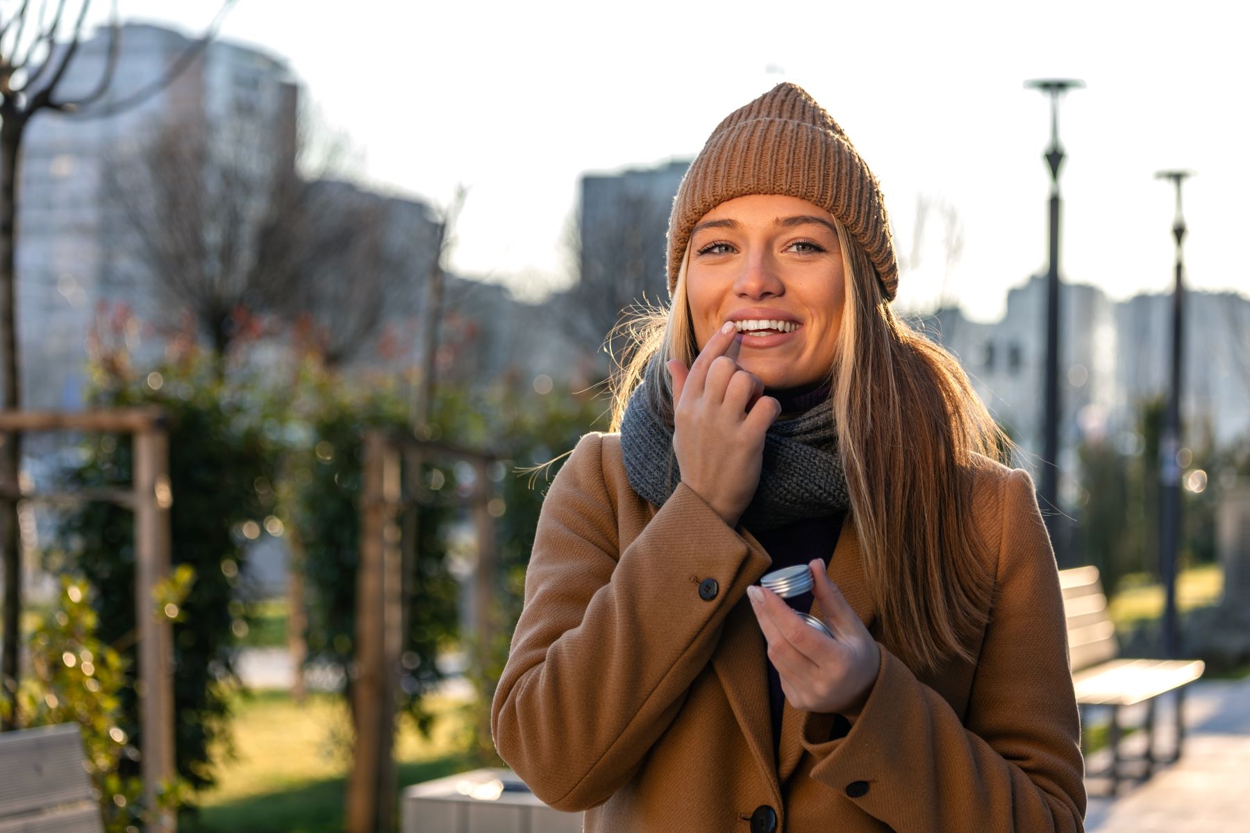Woman applying lip balm outdoors, smiling, wearing a knit hat and coat on a sunny day.