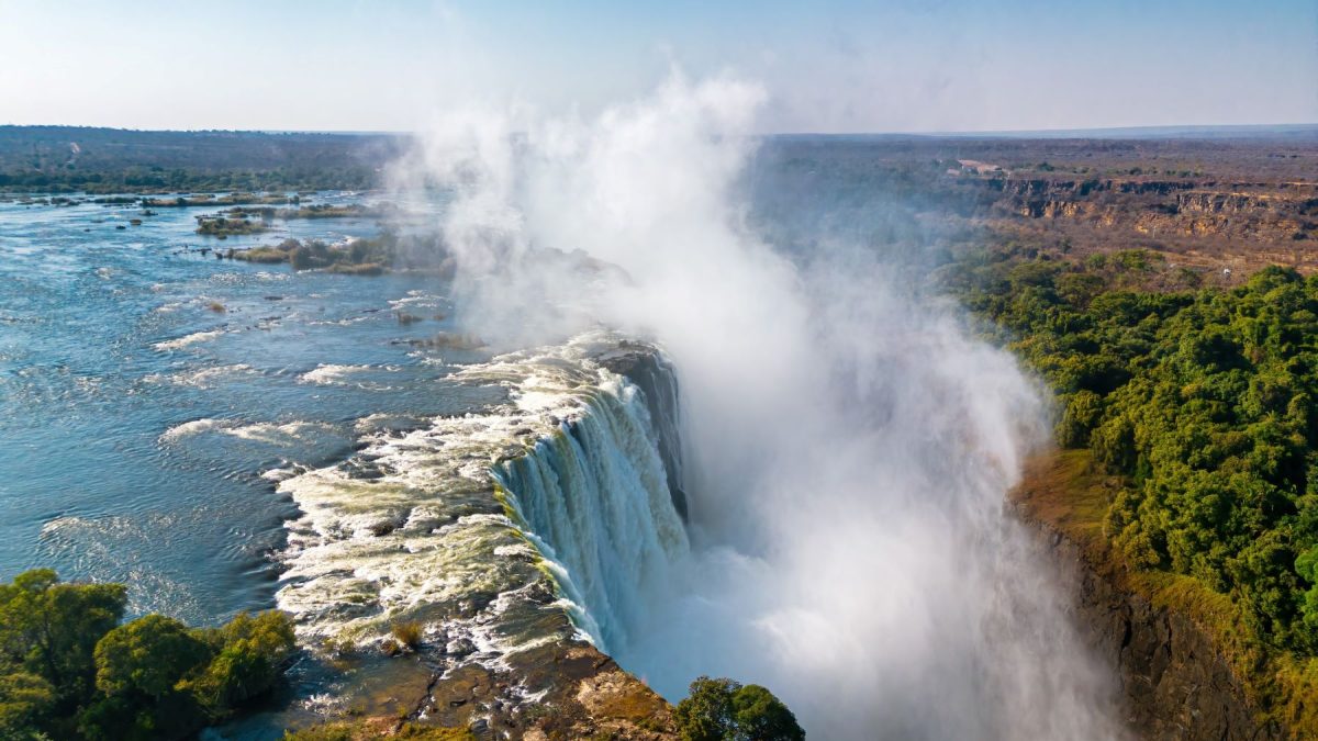 Victoria Falls cascading into the Zambezi River, surrounded by lush greenery under a clear blue sky.