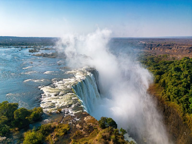 Victoria Falls cascading into the Zambezi River, surrounded by lush greenery under a clear blue sky.