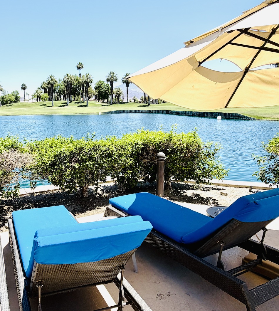 Lounge chairs with blue cushions overlook a serene lake surrounded by palm trees at JW Marriott Desert Springs.