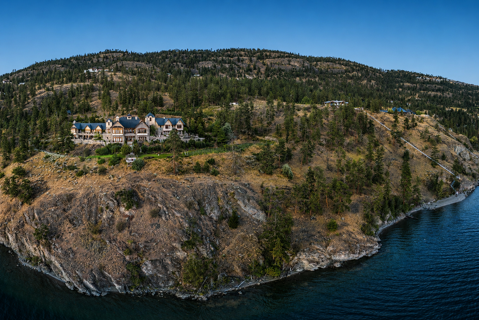 A sprawling lodge sits atop a rocky cliff surrounded by trees, overlooking a serene body of water and distant hills.