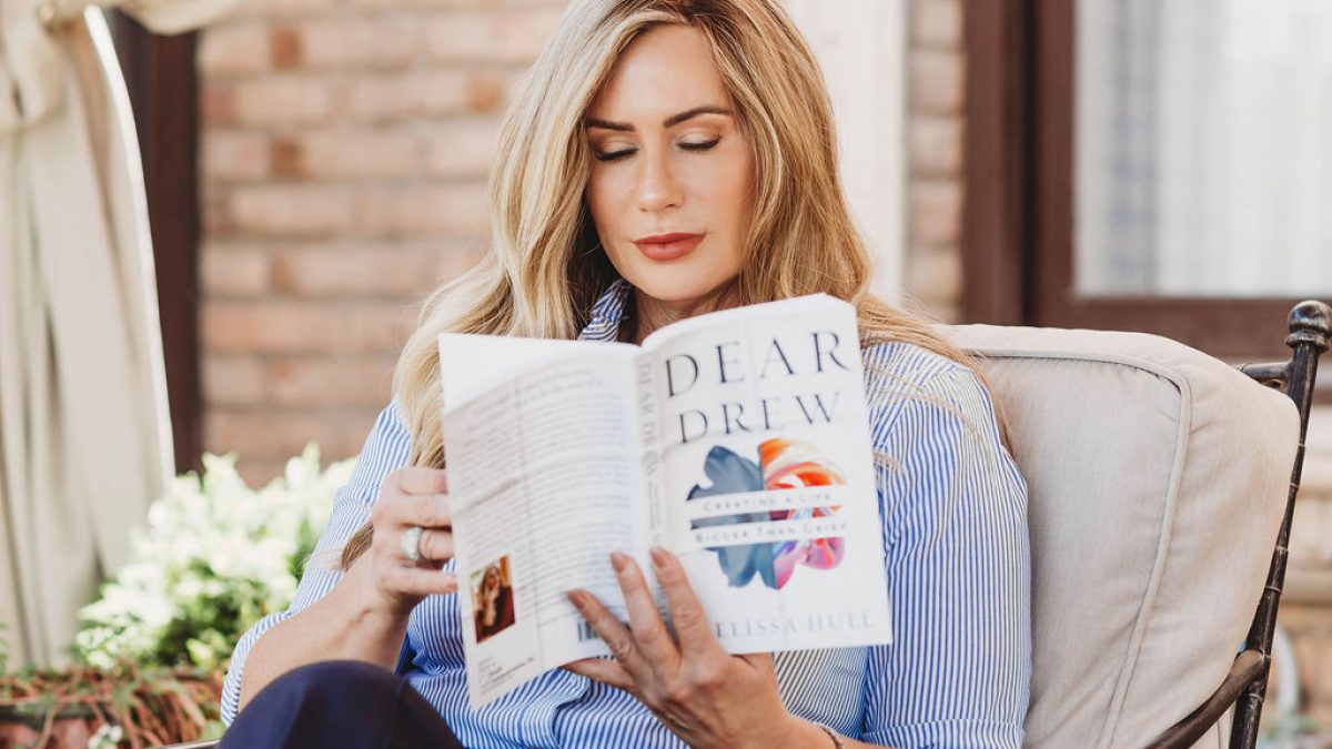 Woman sitting on a patio, reading a book titled "Dear Drew" while relaxing on a comfortable chair.