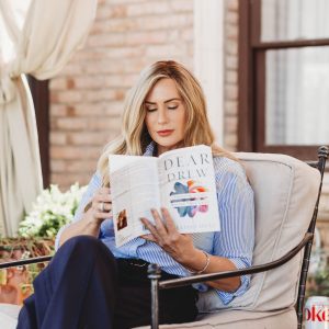 Woman sitting on a patio, reading a book titled "Dear Drew" while relaxing on a comfortable chair.