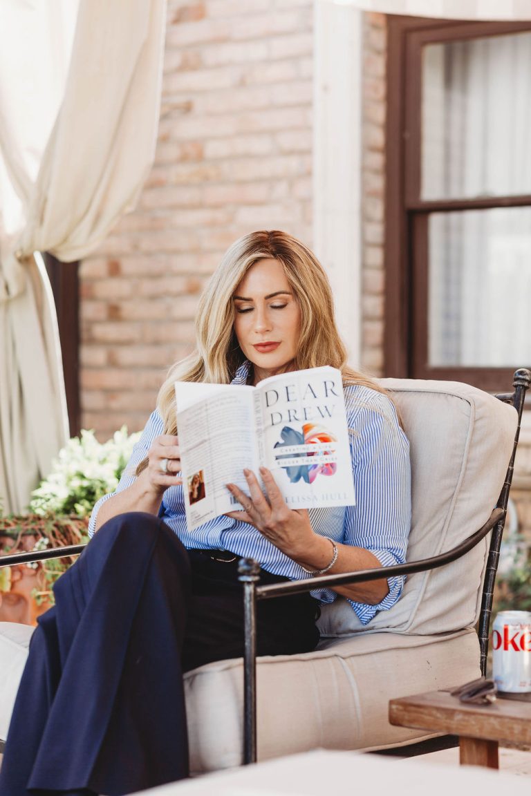 Woman sitting on a patio, reading a book titled "Dear Drew" while relaxing on a comfortable chair.