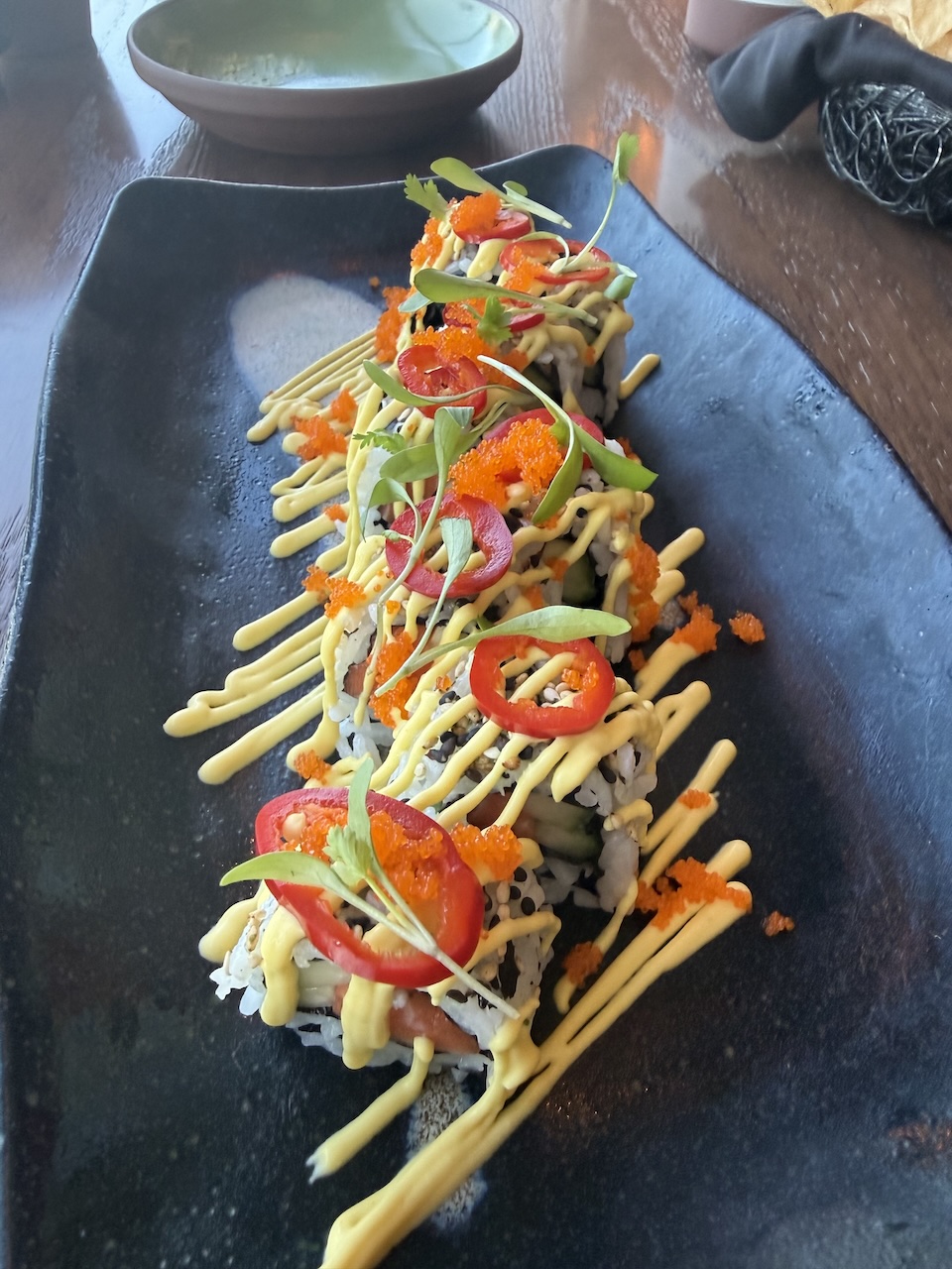 Nikkei sushi rolls topped with spicy mayo, slices of red chili, fish roe, and microgreens on a black plate.