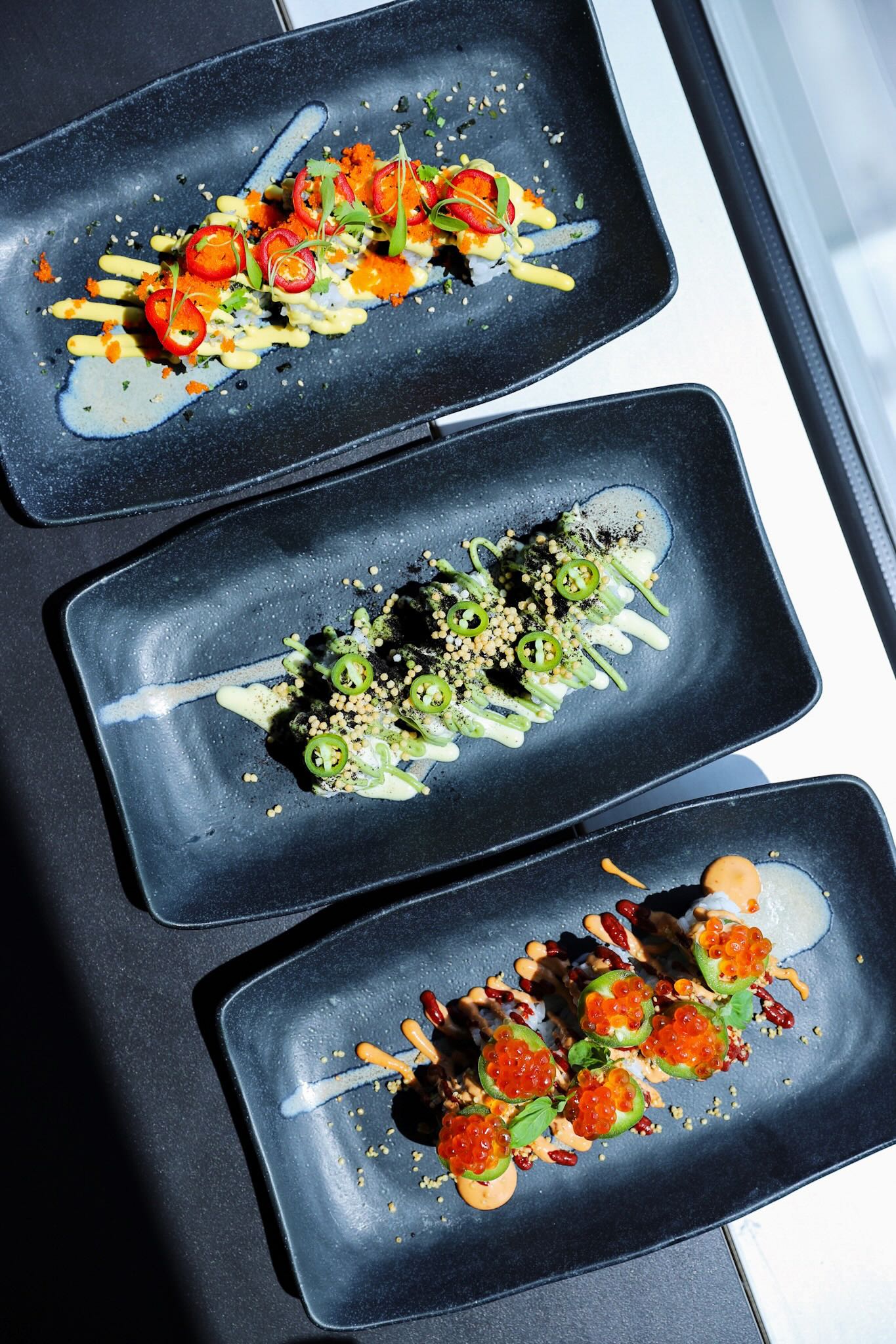 Three elegantly presented sushi dishes with vibrant colors and toppings, served on black rectangular plates.