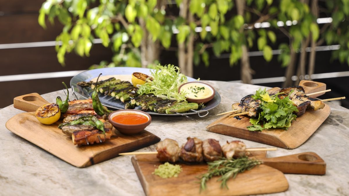 Grilled meats and vegetables served on wooden boards with sauces, garnished with herbs in a lush outdoor setting.
