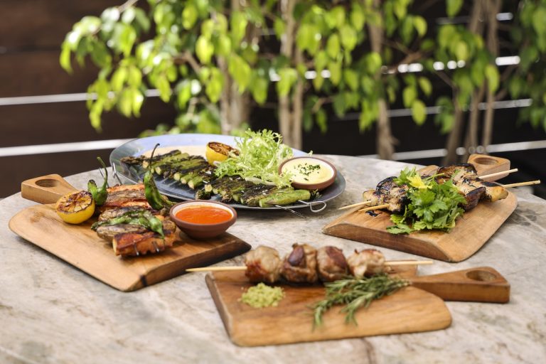 Grilled meats and vegetables served on wooden boards with sauces, garnished with herbs in a lush outdoor setting.