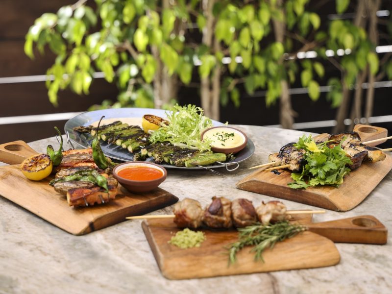 Grilled meats and vegetables served on wooden boards with sauces, garnished with herbs in a lush outdoor setting.