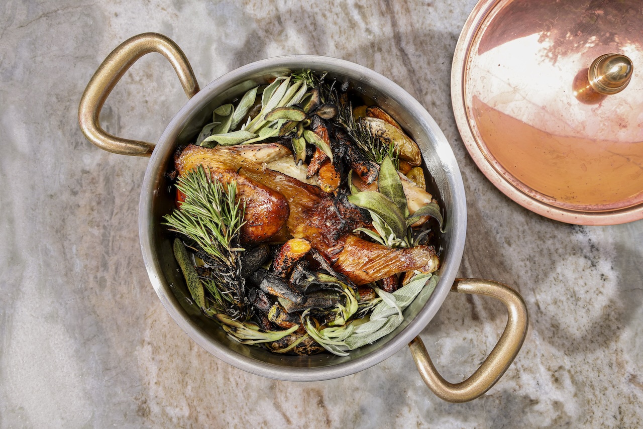 Roasted chicken with herbs and vegetables in a copper pot on a marble surface.