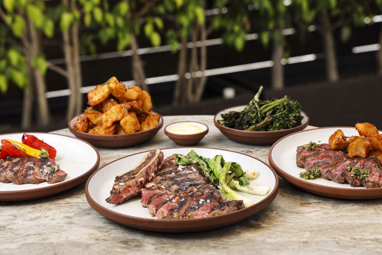 Grilled steak with vegetables and sides, including crispy potatoes and broccolini, presented on rustic plates.