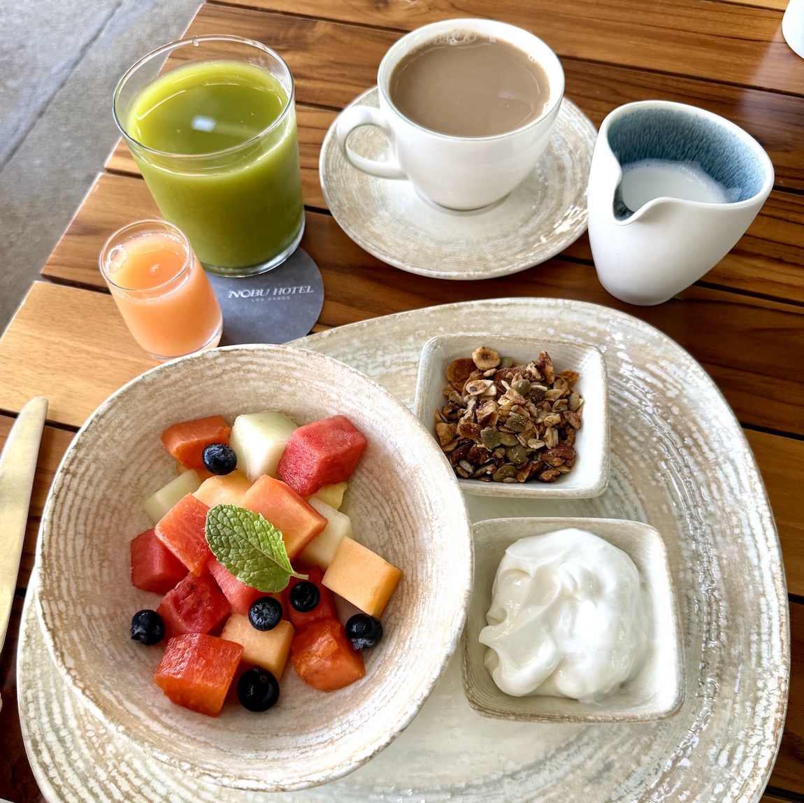 A nutritious breakfast featuring fresh fruit salad, yogurt, granola, a green smoothie, coffee, and a small juice shot.