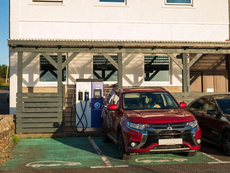 Red electric vehicle parked near a charging station outside a building, illustrating modern charging habits for staycations.