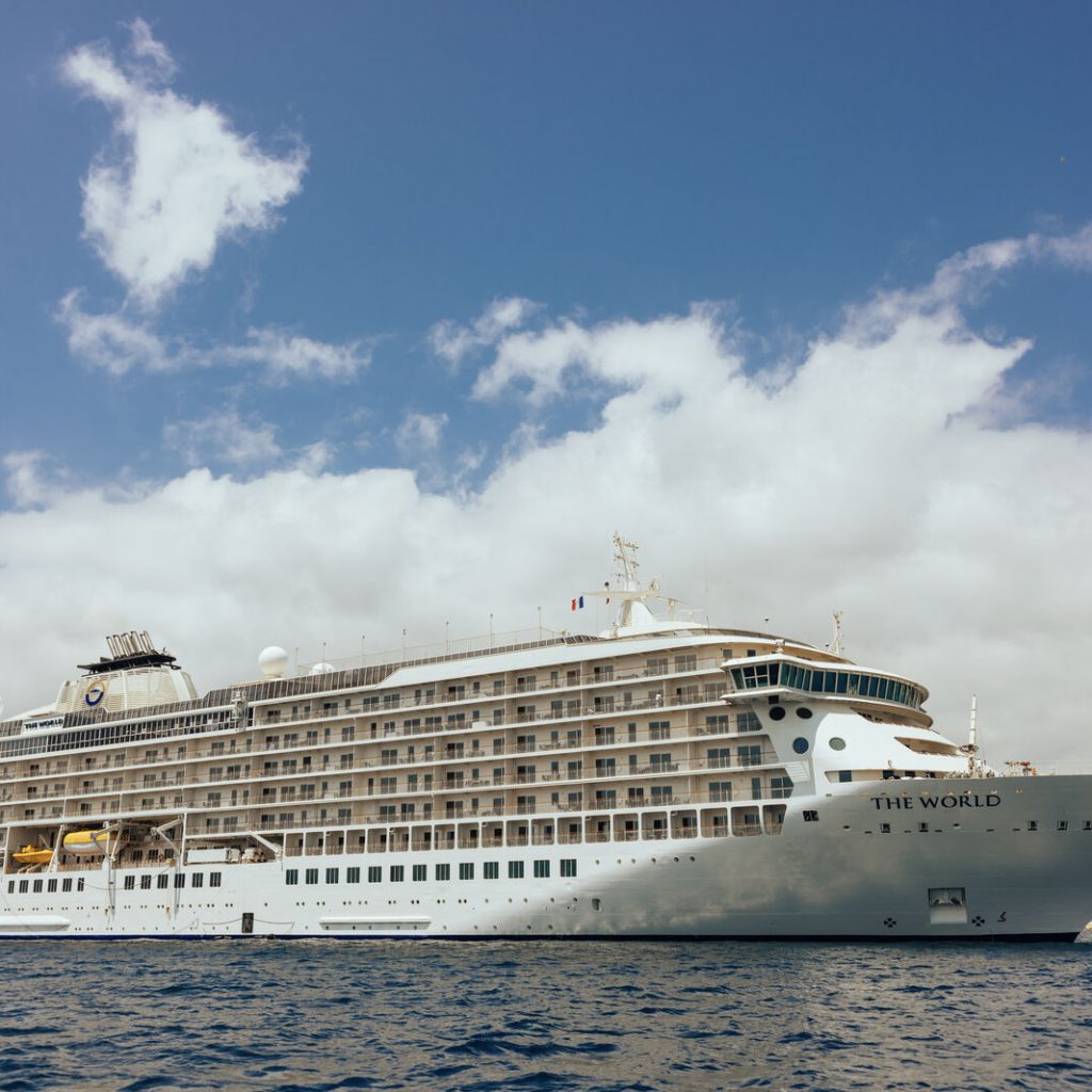 Luxury cruise ship The World anchored in calm waters under a blue sky with clouds.