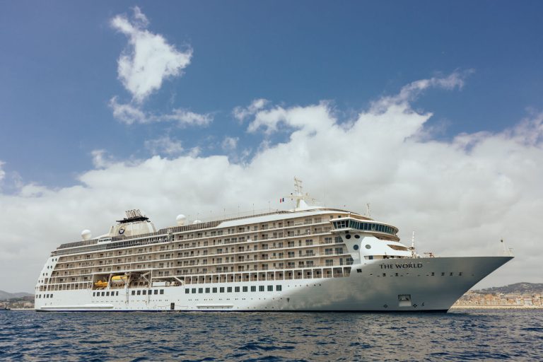 Luxury cruise ship The World anchored in calm waters under a blue sky with clouds.