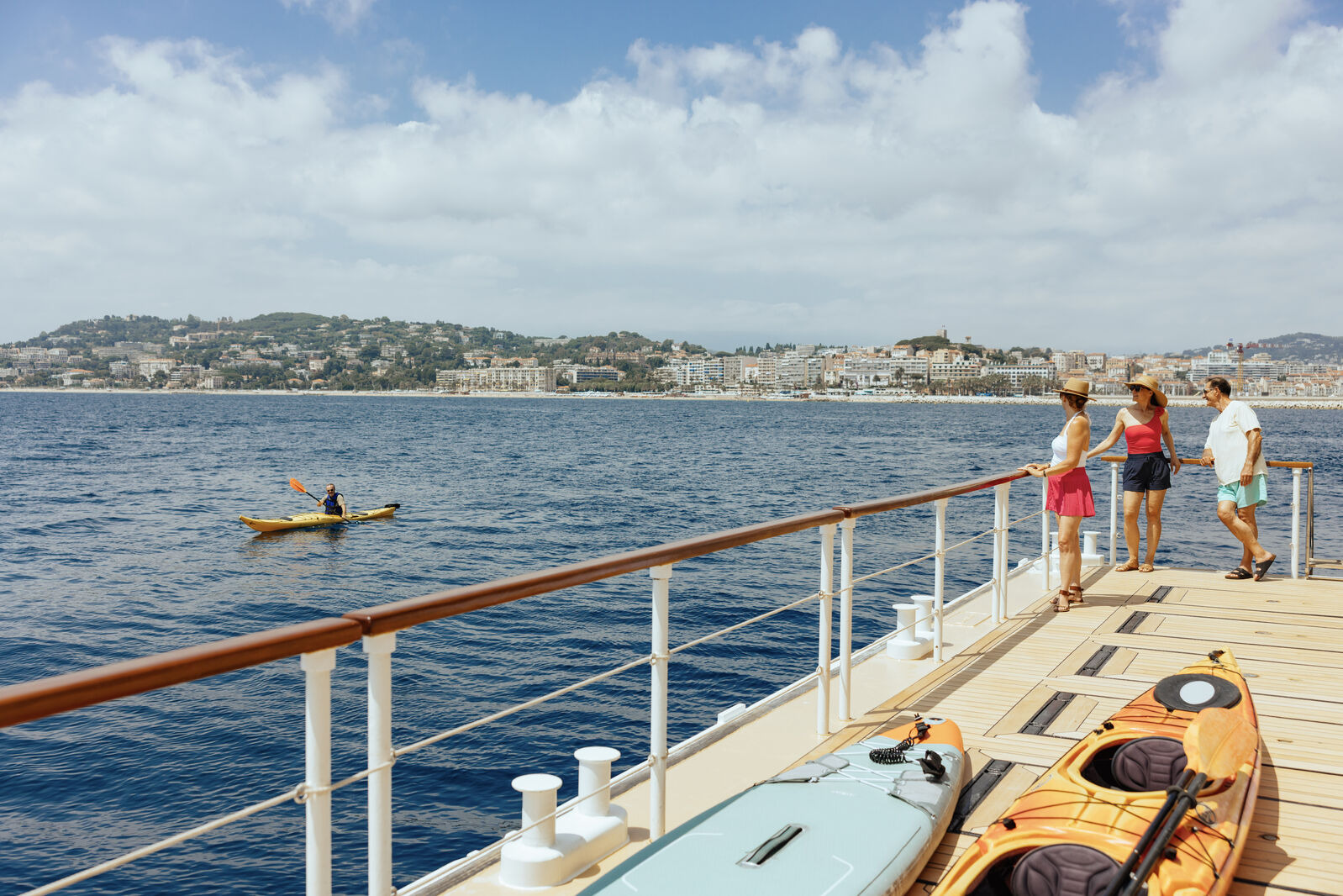Three people enjoy the view from the deck of a luxury ship, while a kayaker paddles in the sea with a coastal city in the background.