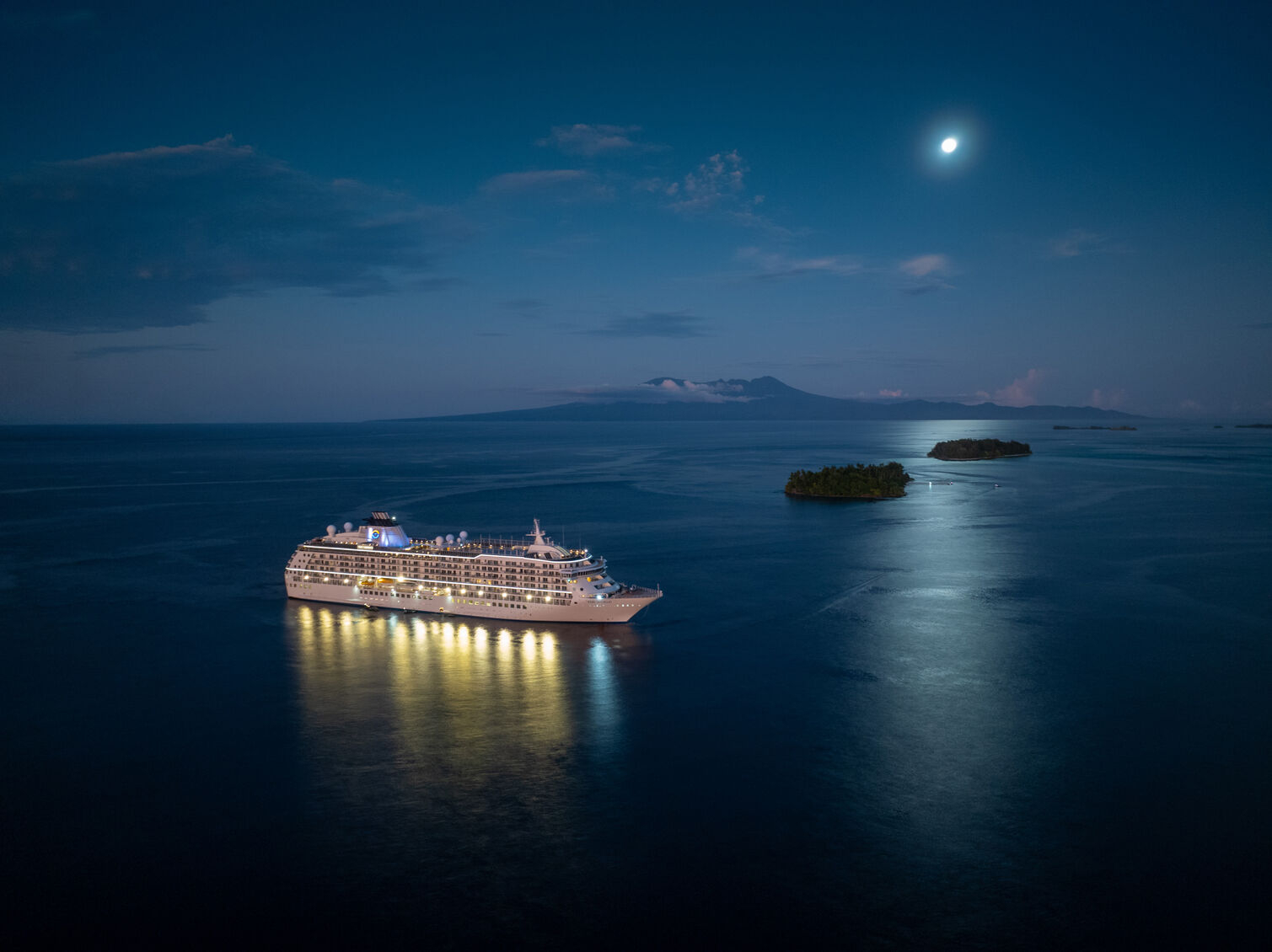 Luxury ship illuminated at night sailing through calm waters near small islands under a moonlit sky.