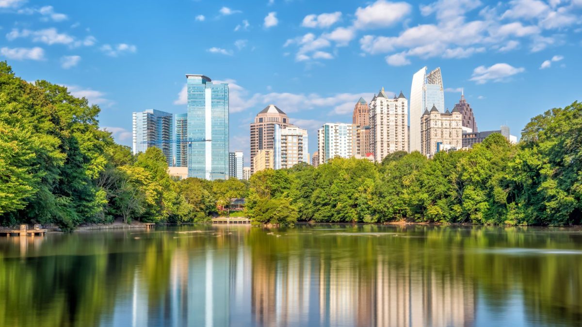 Atlanta skyline reflecting in a lake, surrounded by lush greenery, under a clear blue sky with scattered clouds.