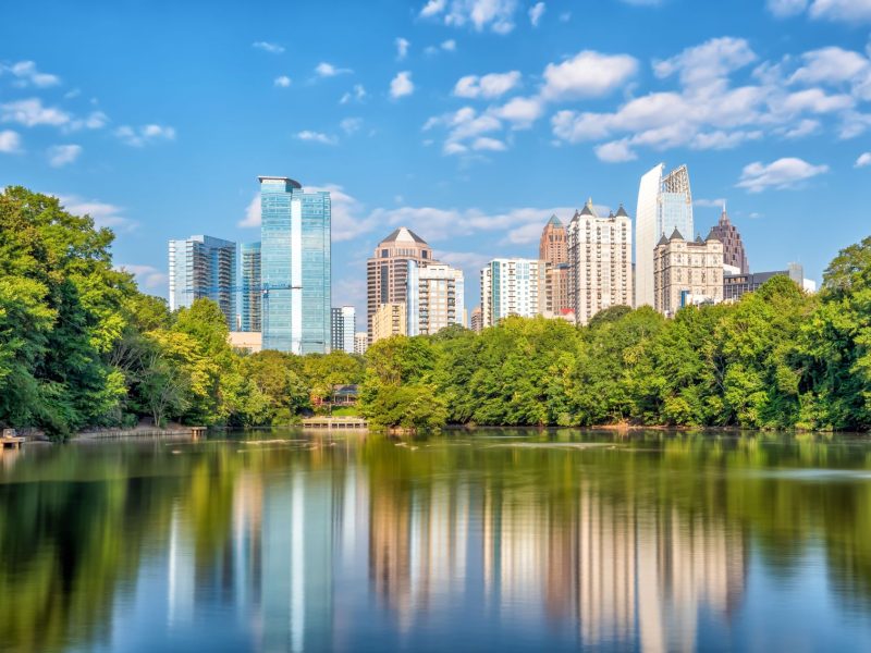 Atlanta skyline reflecting in a lake, surrounded by lush greenery, under a clear blue sky with scattered clouds.