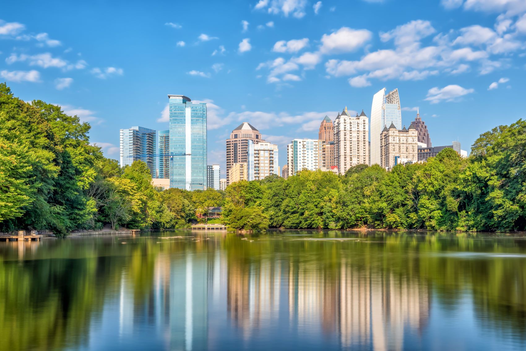 Atlanta skyline reflecting in a lake, surrounded by lush greenery, under a clear blue sky with scattered clouds.