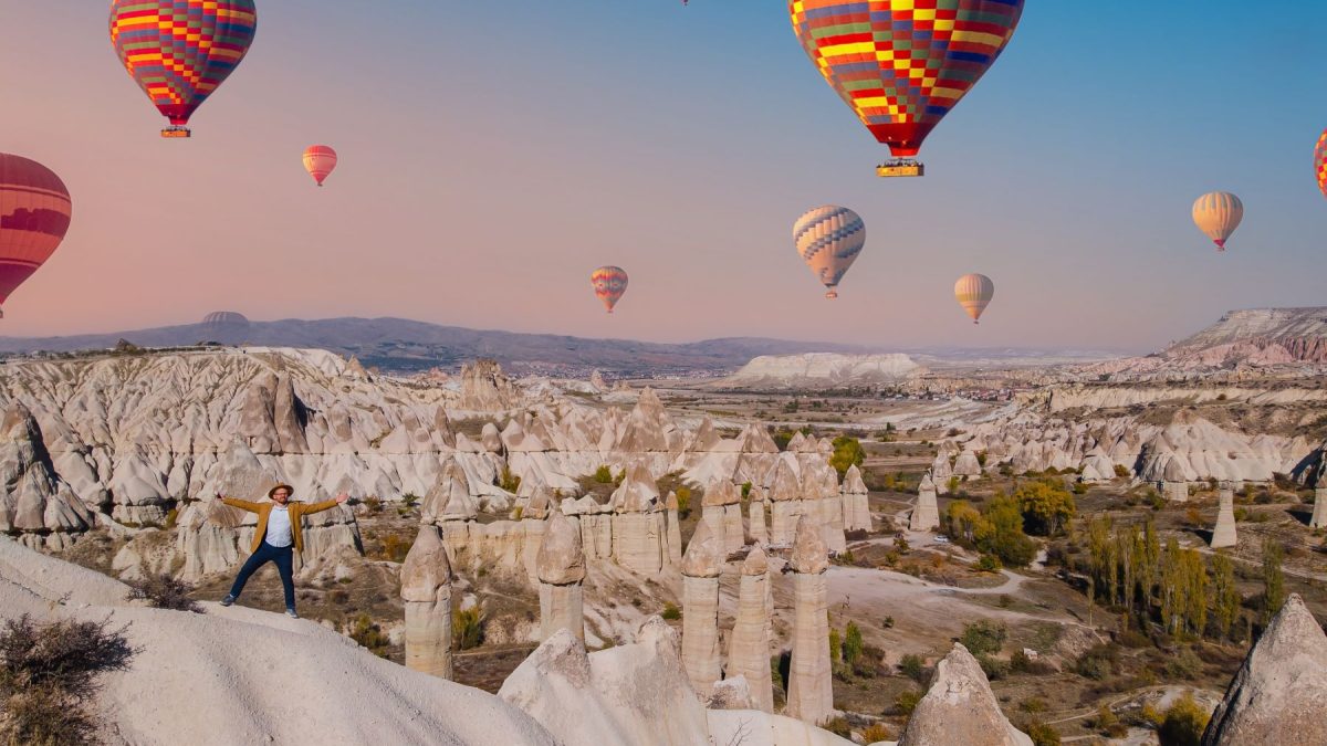 Hot air balloons soar above unique rock formations as a person joyfully stands on a ridge in a picturesque landscape.