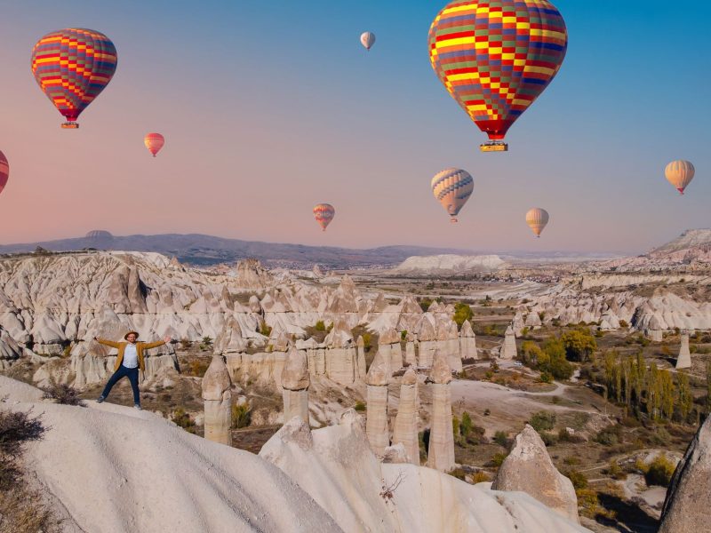 Hot air balloons soar above unique rock formations as a person joyfully stands on a ridge in a picturesque landscape.