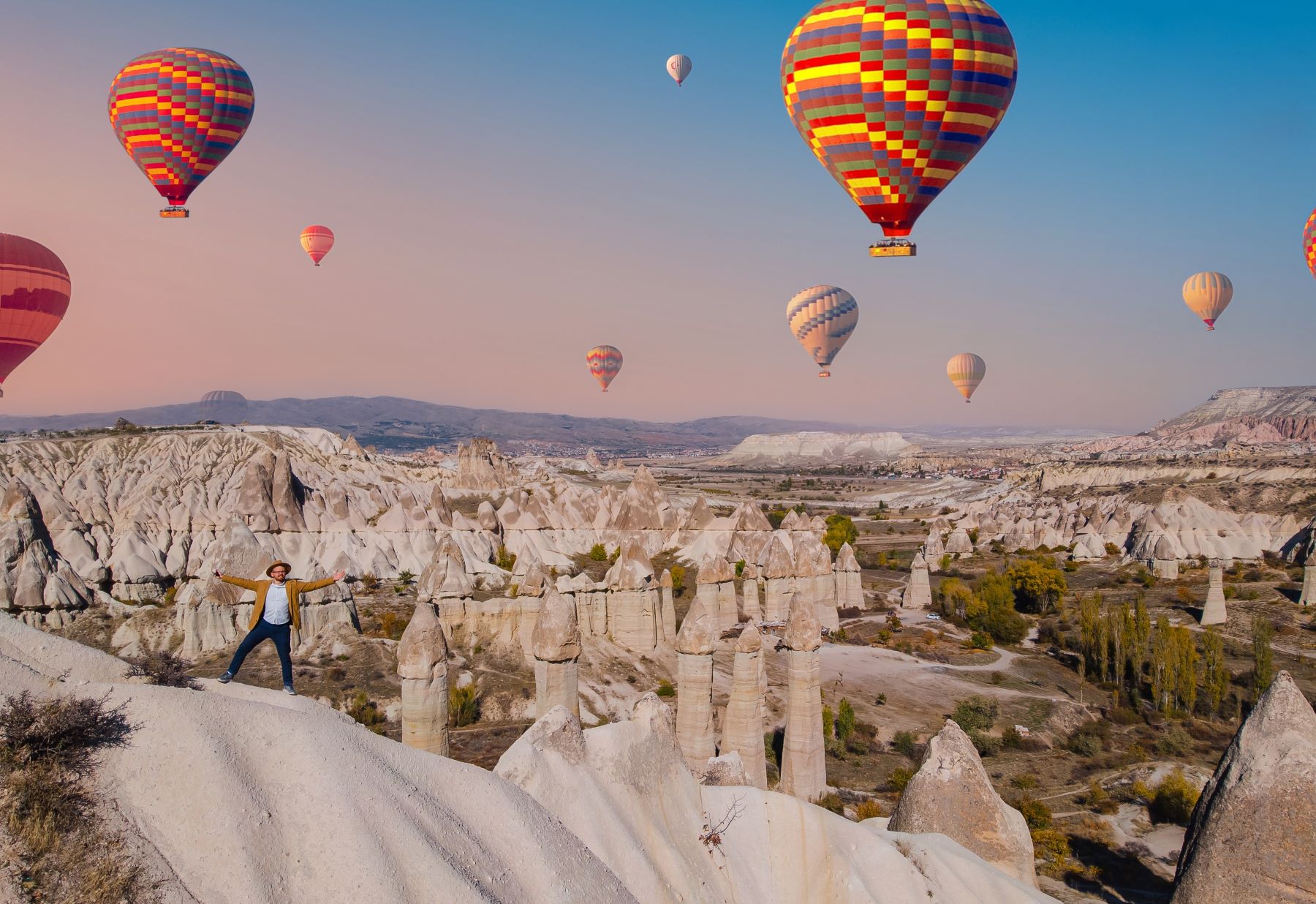 Hot air balloons soar above unique rock formations as a person joyfully stands on a ridge in a picturesque landscape.