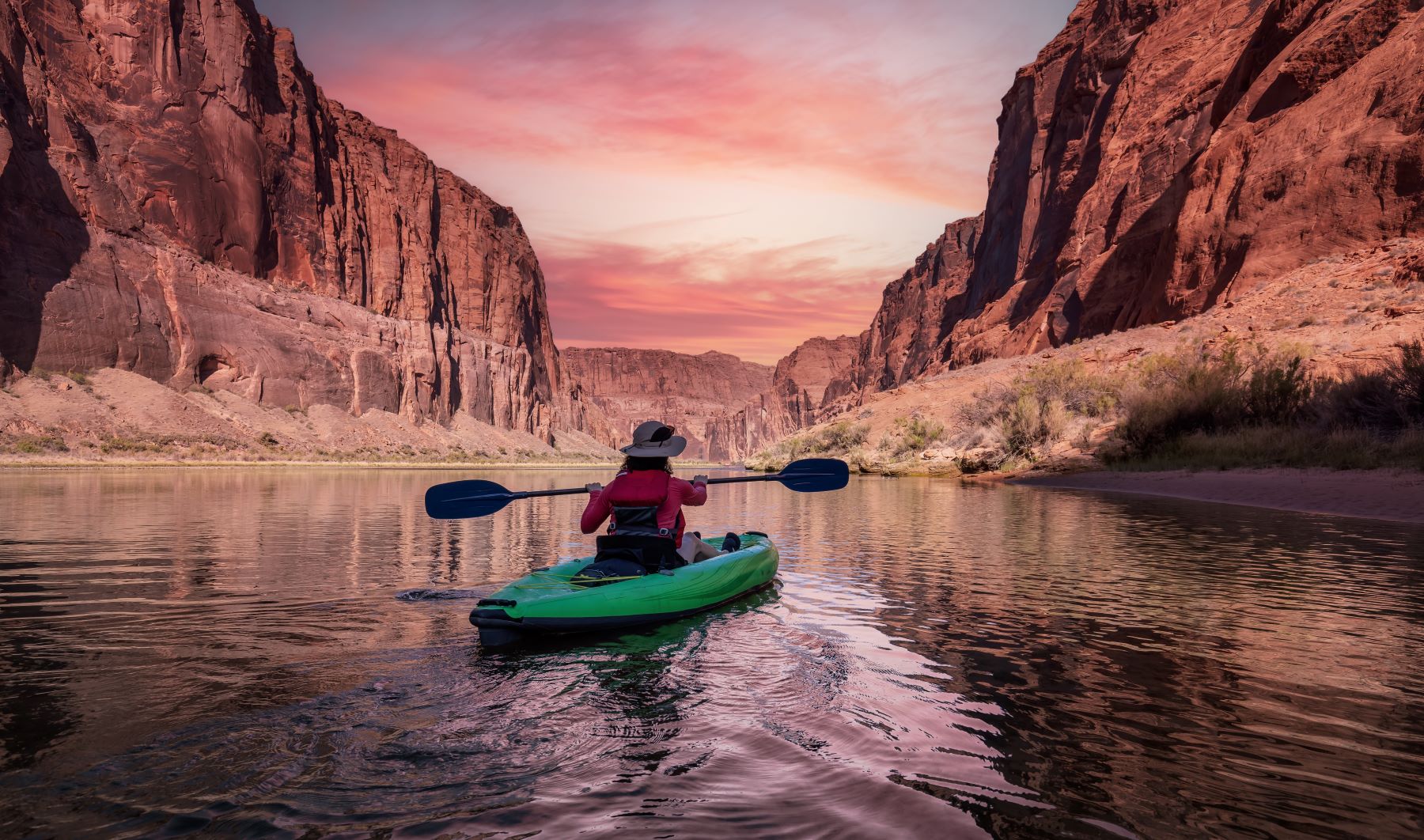 Kayaker paddling through a serene canyon at sunset, surrounded by towering red rock cliffs and reflecting water.