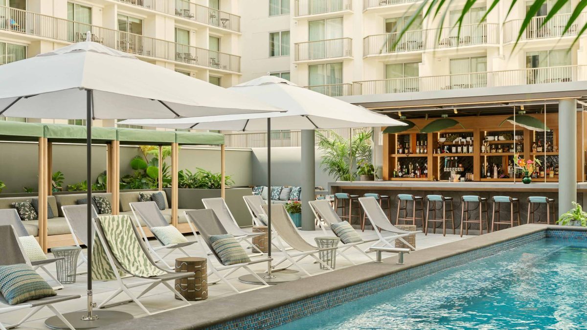 Lounge chairs and umbrellas surround a pool, with a bar and greenery enhancing the inviting atmosphere of the backyard.
