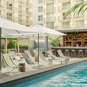 Lounge chairs and umbrellas surround a pool, with a bar and greenery enhancing the inviting atmosphere of the backyard.