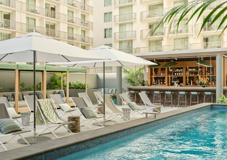 Lounge chairs and umbrellas surround a pool, with a bar and greenery enhancing the inviting atmosphere of the backyard.