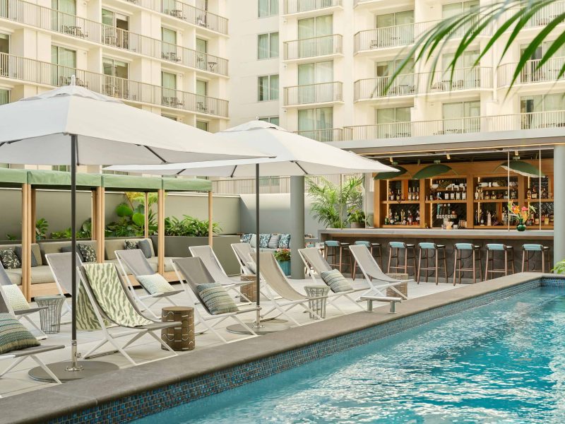 Lounge chairs and umbrellas surround a pool, with a bar and greenery enhancing the inviting atmosphere of the backyard.