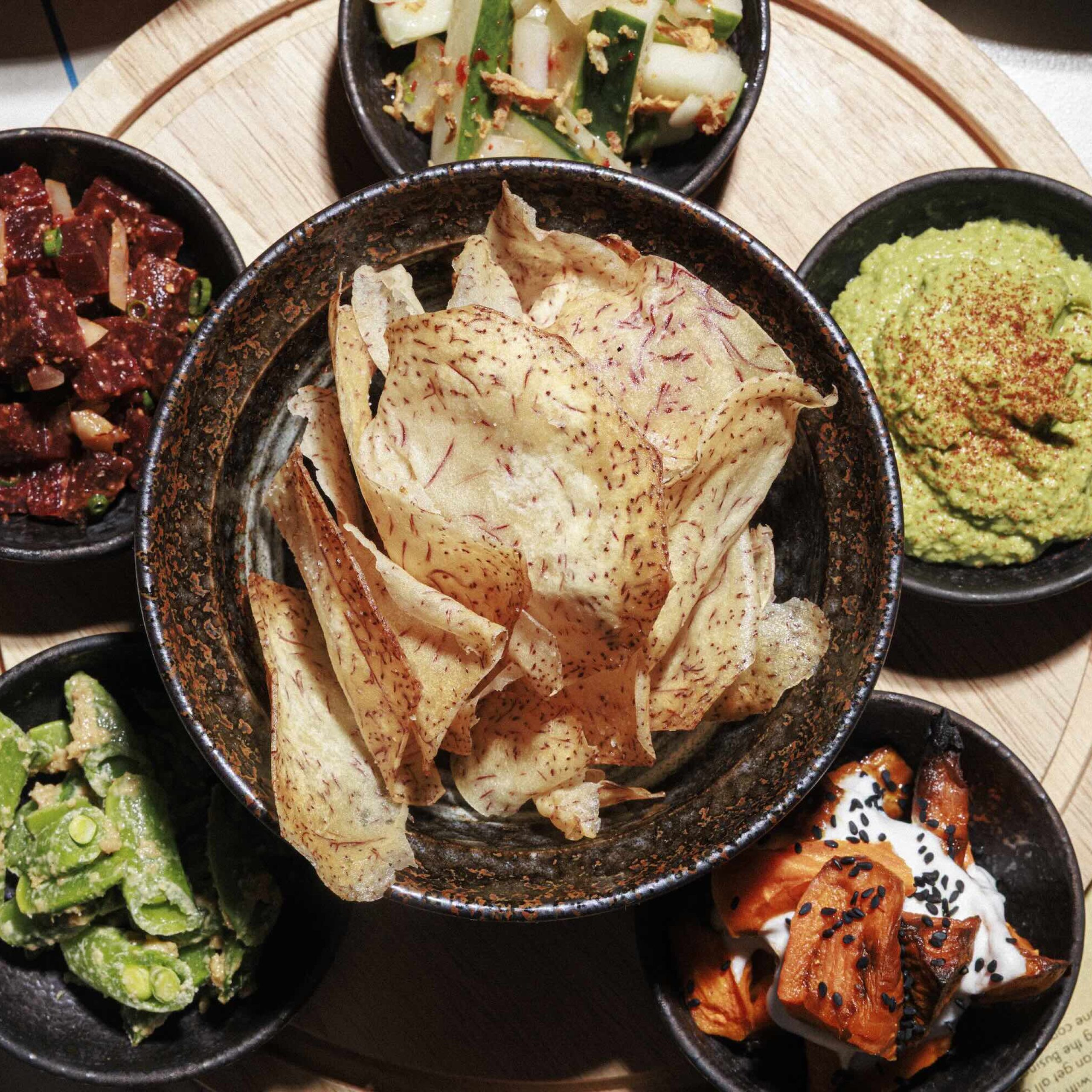 A platter featuring various dips and snacks, including taro chips, poke, and salads served in black bowls.