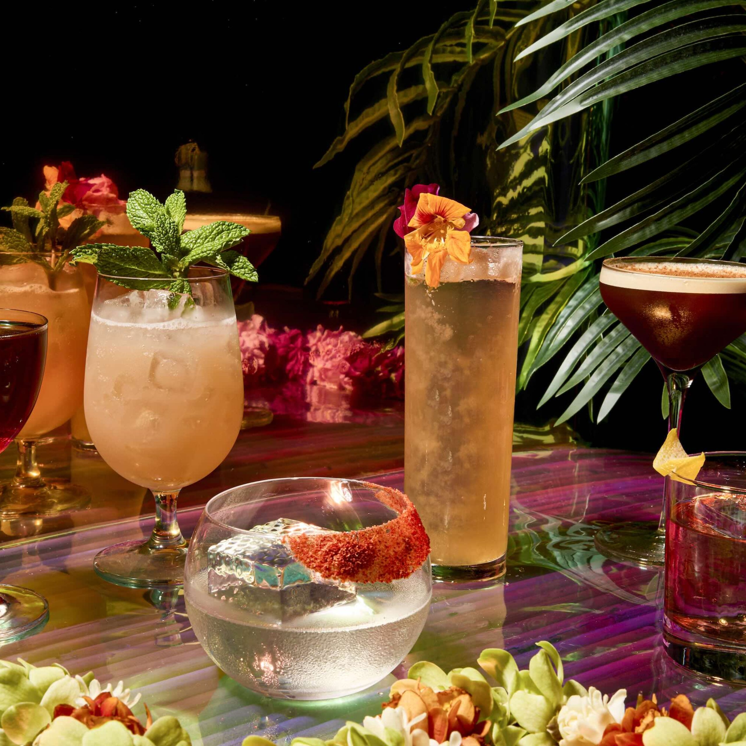 Colorful cocktails arranged on a table surrounded by tropical flowers and greenery.