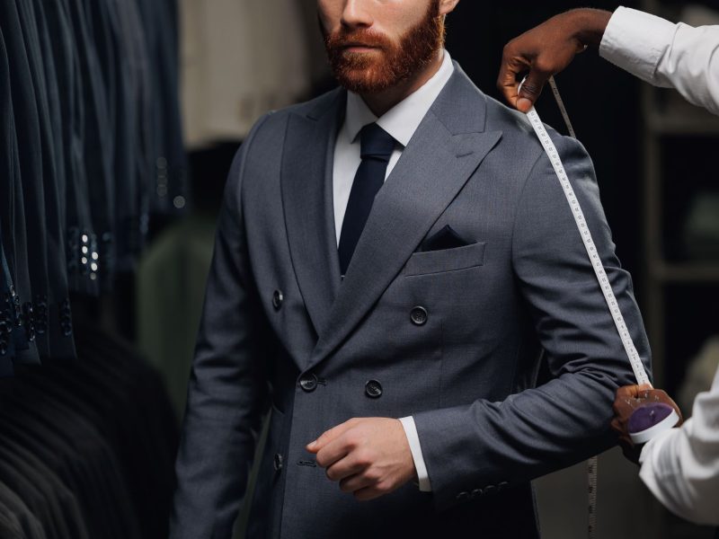 Tailor measuring a man's sleeve while he wears a stylish grey suit, surrounded by various suits in the background.