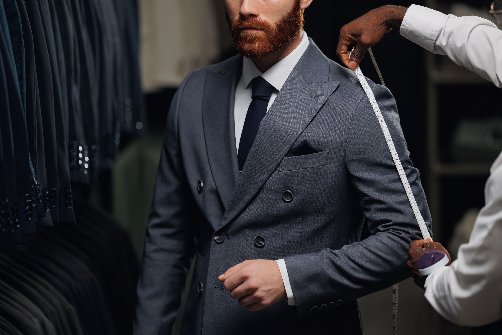 Tailor measuring a man's sleeve while he wears a stylish grey suit, surrounded by various suits in the background.