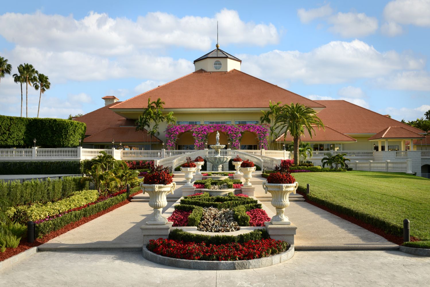 Luxurious resort entrance featuring a grand fountain, vibrant flowers, and palm trees under a clear blue sky.