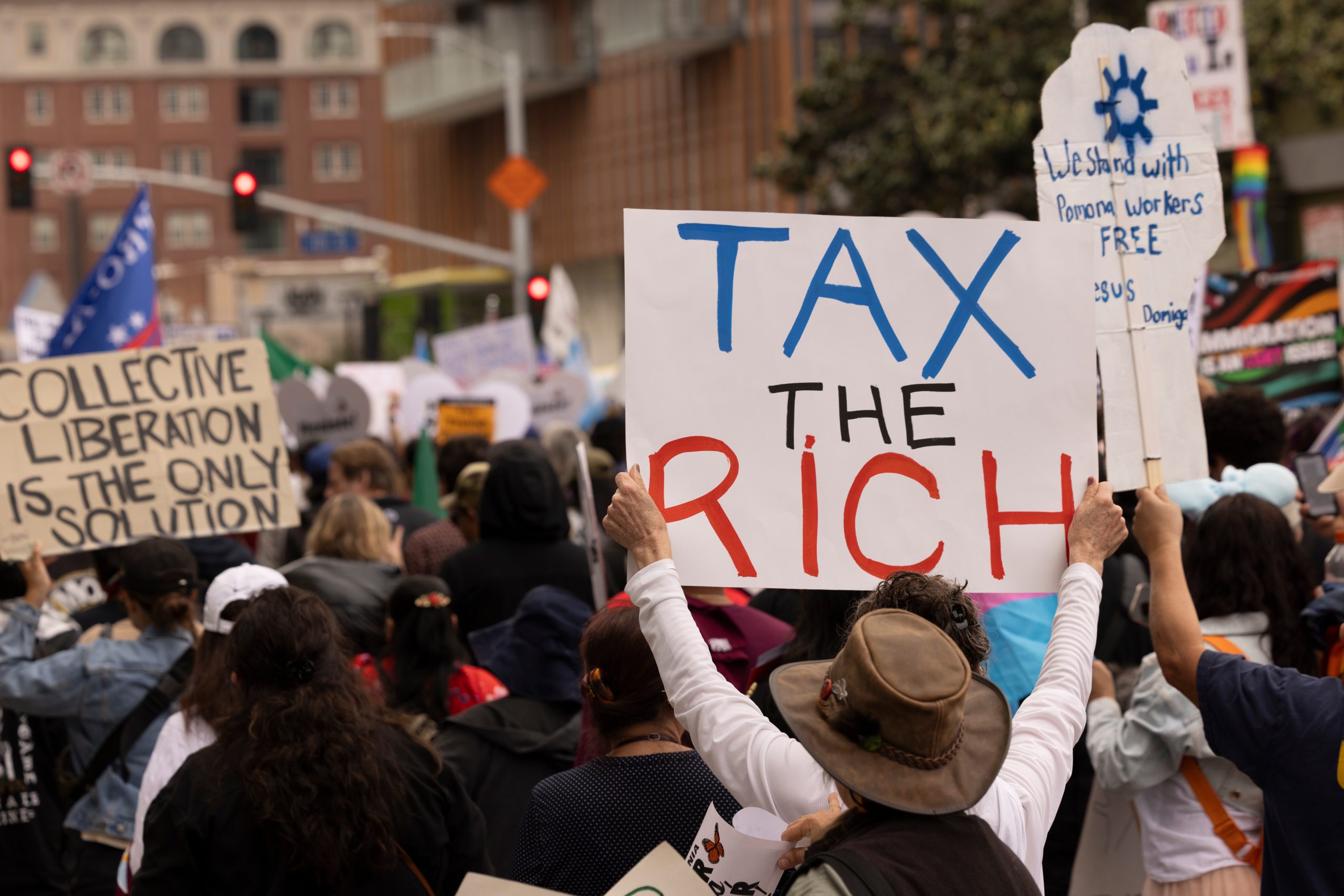 Crowd at a protest holding signs advocating for wealth redistribution and economic justice, including "Tax the rich."