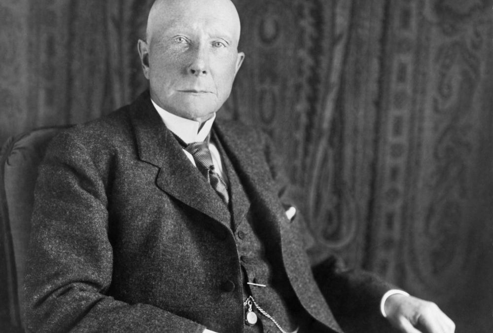 Older man in formal attire, seated with a serious expression against a patterned backdrop.