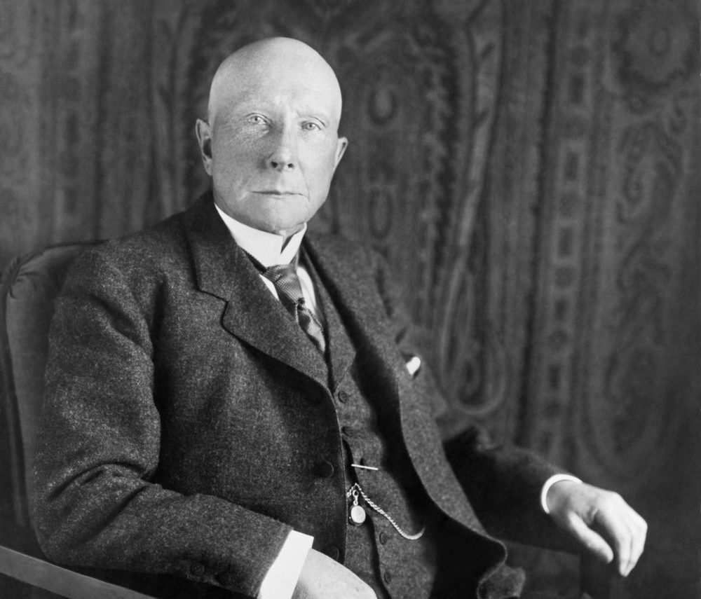 Older man in formal attire, seated with a serious expression against a patterned backdrop.