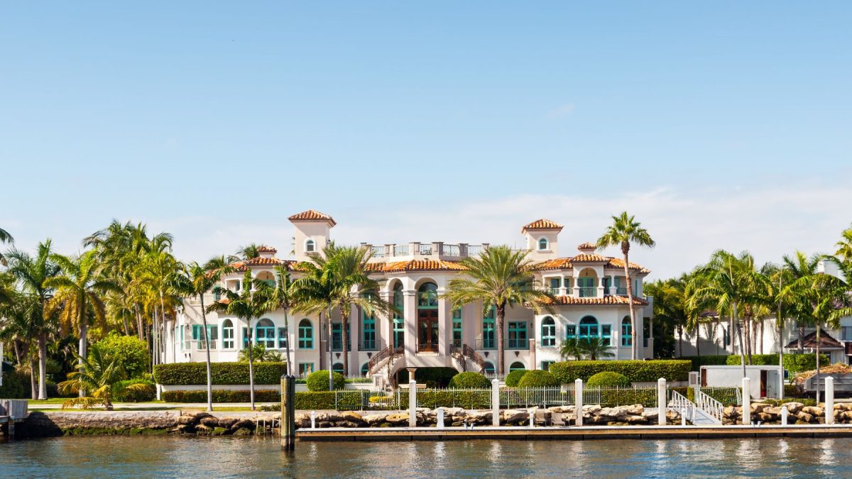 Luxurious waterfront mansion surrounded by palm trees, with a clear blue sky in the background. Perfect for luxury real estate inspiration.