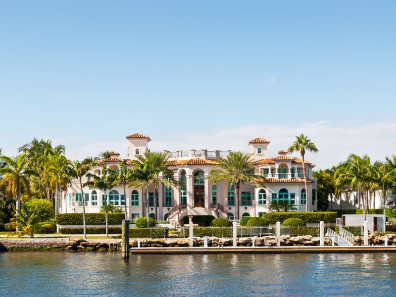 Luxurious waterfront mansion surrounded by palm trees, with a clear blue sky in the background. Perfect for luxury real estate inspiration.