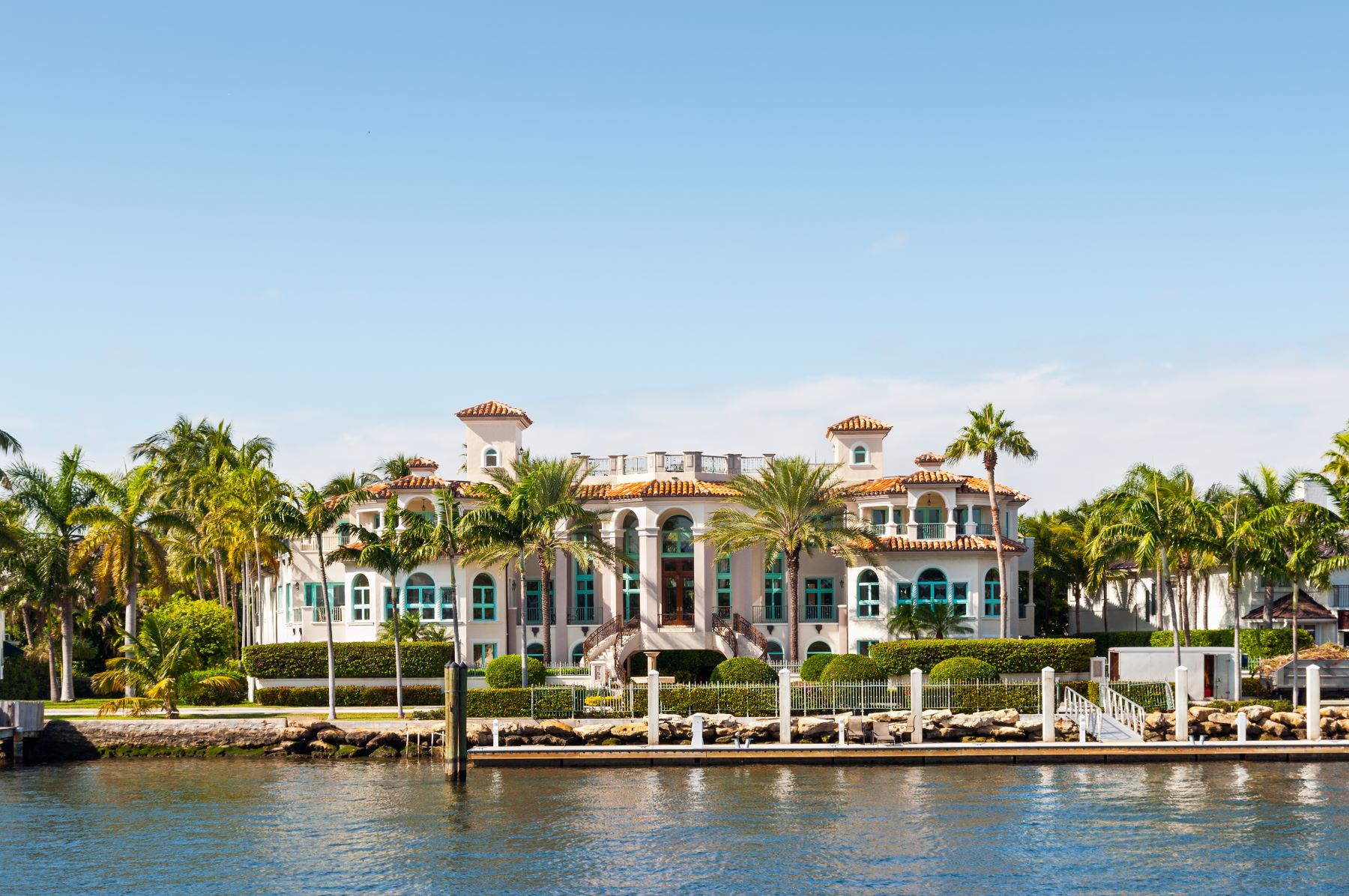 Luxurious waterfront mansion surrounded by palm trees, with a clear blue sky in the background. Perfect for luxury real estate inspiration.