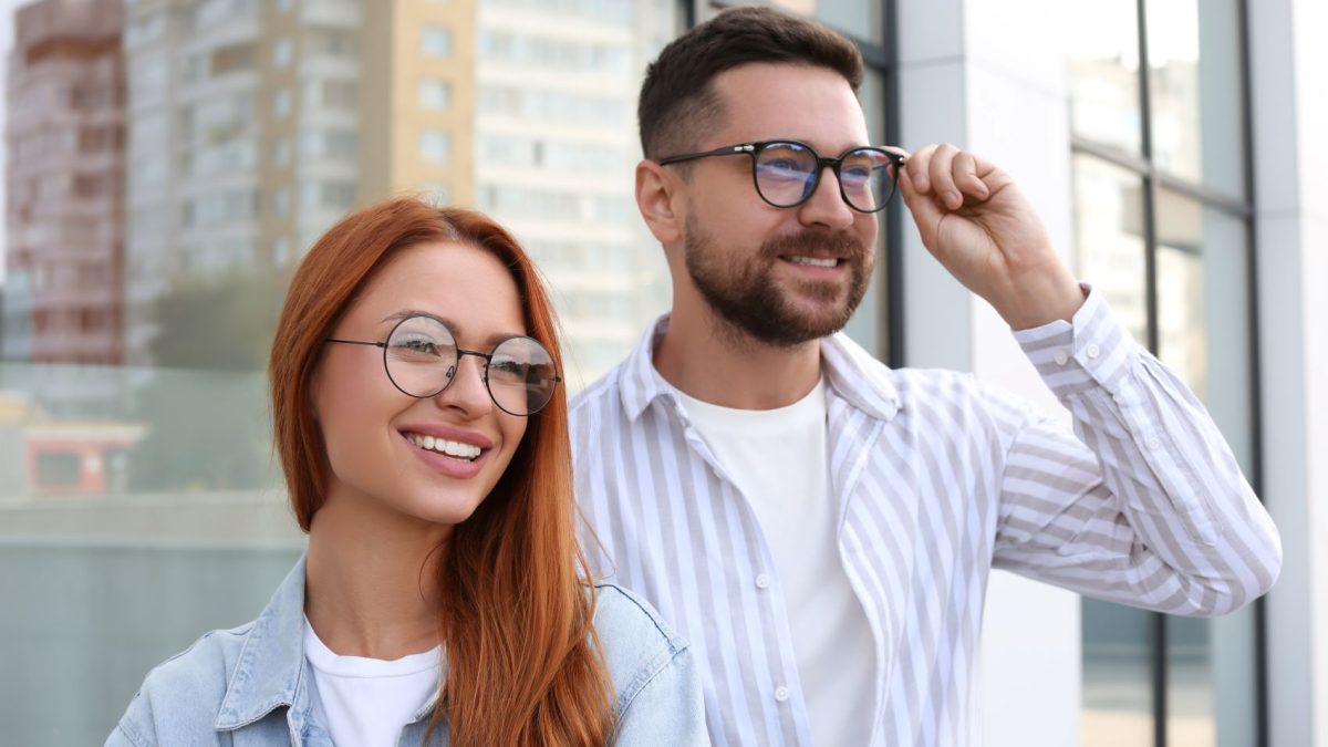 A smiling woman with long red hair and a man in glasses pose together outside a modern building, showcasing stylish eyewear.