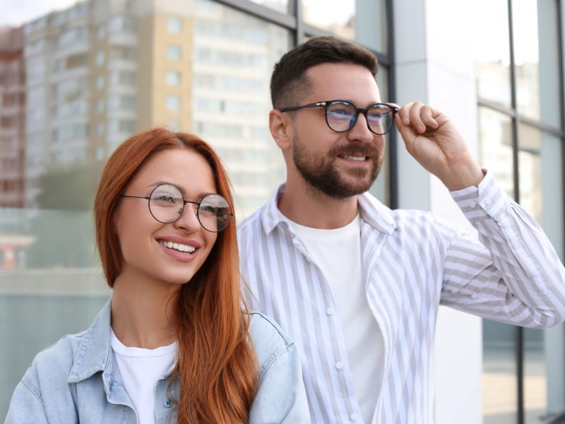 A smiling woman with long red hair and a man in glasses pose together outside a modern building, showcasing stylish eyewear.