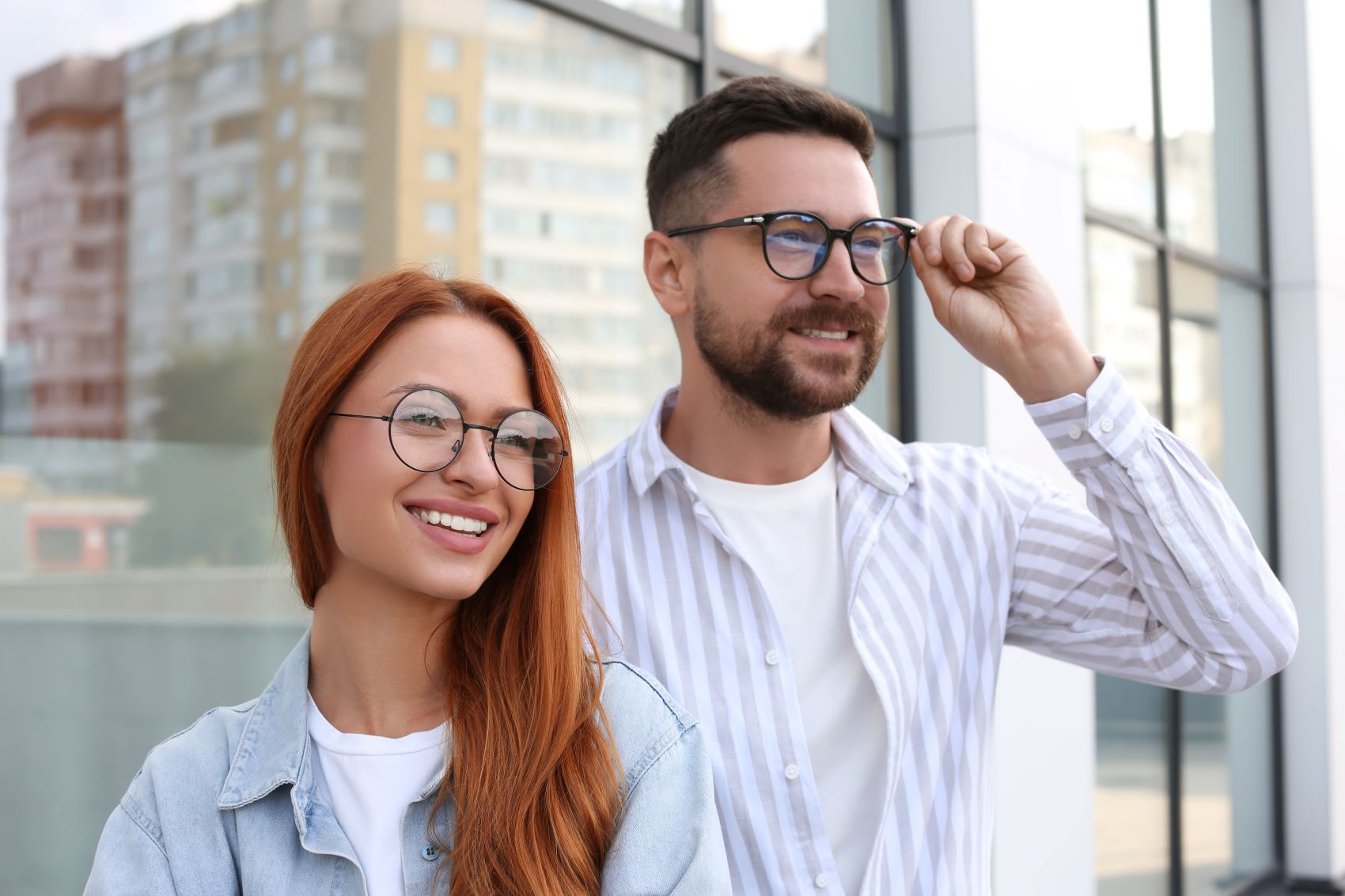 A smiling woman with long red hair and a man in glasses pose together outside a modern building, showcasing stylish eyewear.