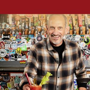 Entrepreneur holding a Bloody Mary, smiling in front of a bar adorned with stickers and beer taps.